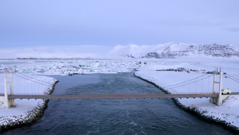 AERIAL:-Flying-over-Bridge-with-Car-driving-over-towards-Snowy-Ice-Floes-on-Iceland-Lake-Winter,-Snow