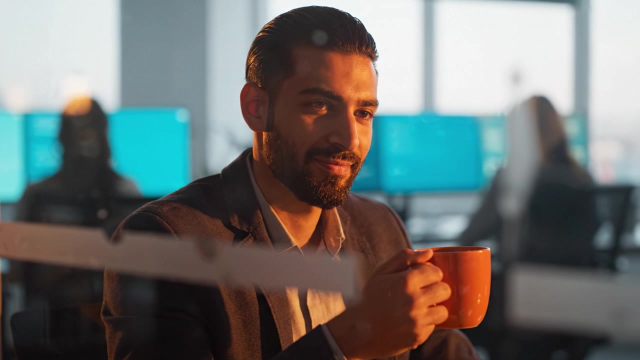 Un momento contemplativo catturato in uno spazio di lavoro moderno, che mostra un uomo che si gode una bevanda calda mentre riflette e sorride in un vivace ambiente d'ufficio.