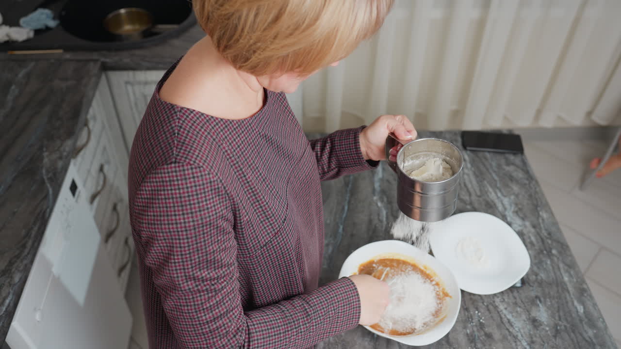 воздушный вид женщины, смешивающей густой суп в белой чаше, когда в ней просыпают мелкую белую кукурузную муку, виск плавно смешивает ингредиенты, смартфон лежит на темной столешнице