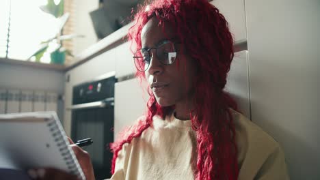 niña afroamericana con cabello rizado rojo estudiando en el suelo de la cocina, tomando notas