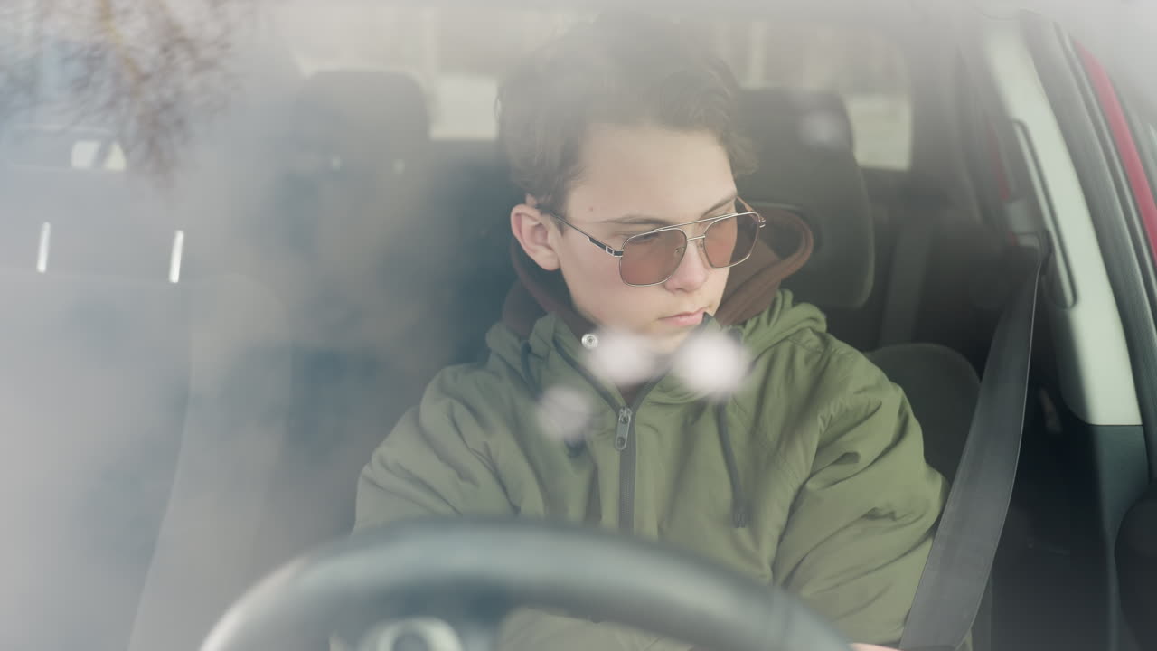student seen through windshield removing seatbelt inside parked vehicle during snowy winter day, wearing green puffy jacket and sunglasses, faint snowflakes and reflections visible on glass surface