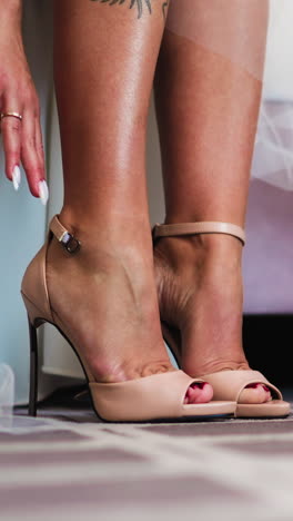 young bride in white dress puts on and clasps elegant shoes with high stiletto heels sitting on bed at home close view slow motion