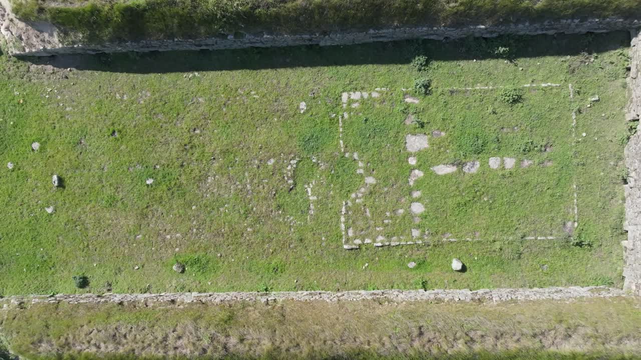 Drone ascending in top-down view over an 18th-century stone structure for pig farming. Visible feeding and watering trough remains inside. Surrounded by open countryside landscape.