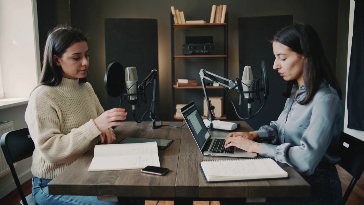 Donna a sinistra che esamina le pagine e prende il microfono per avviare un podcast in una stanza, mentre il co-conduttore digita al laptop