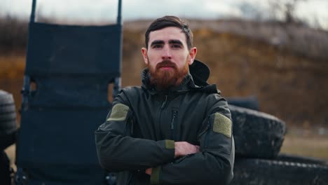 Portrait-of-a-confident-brunette-man-with-a-beard-in-a-dark-green-army-uniform-near-barricades-made-of-rubber-tires-of-a-car-outside-the-city