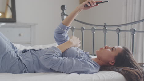 woman wearing pyjamas posing and taking selfies on mobile phone lying on bed