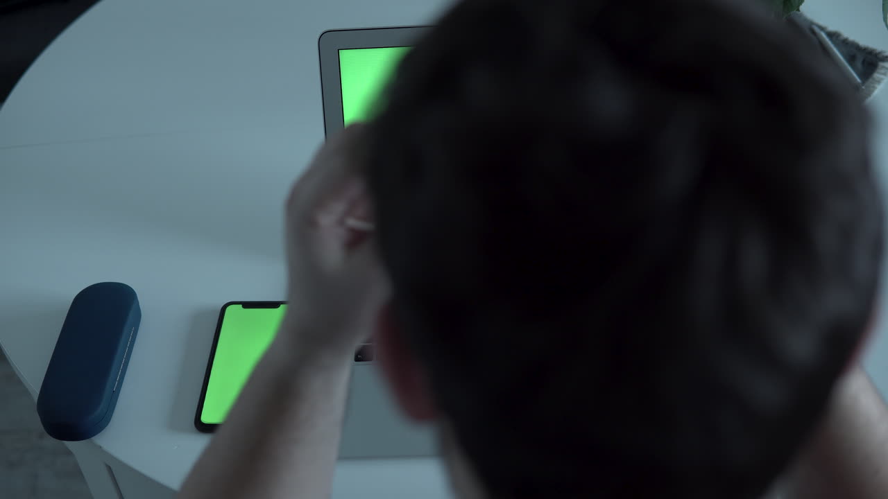 Green screen on mobile phone and laptop monitor, top view man sitting neat table and take glasses, home office