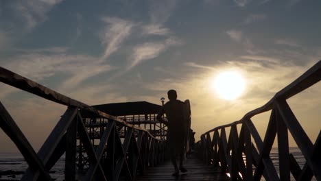 Eine-Kamerafahrt-In-Zeitlupe-Eines-Männlichen-Surfers,-Der-In-Der-Abenddämmerung-Den-Pier-Hinunterjoggt