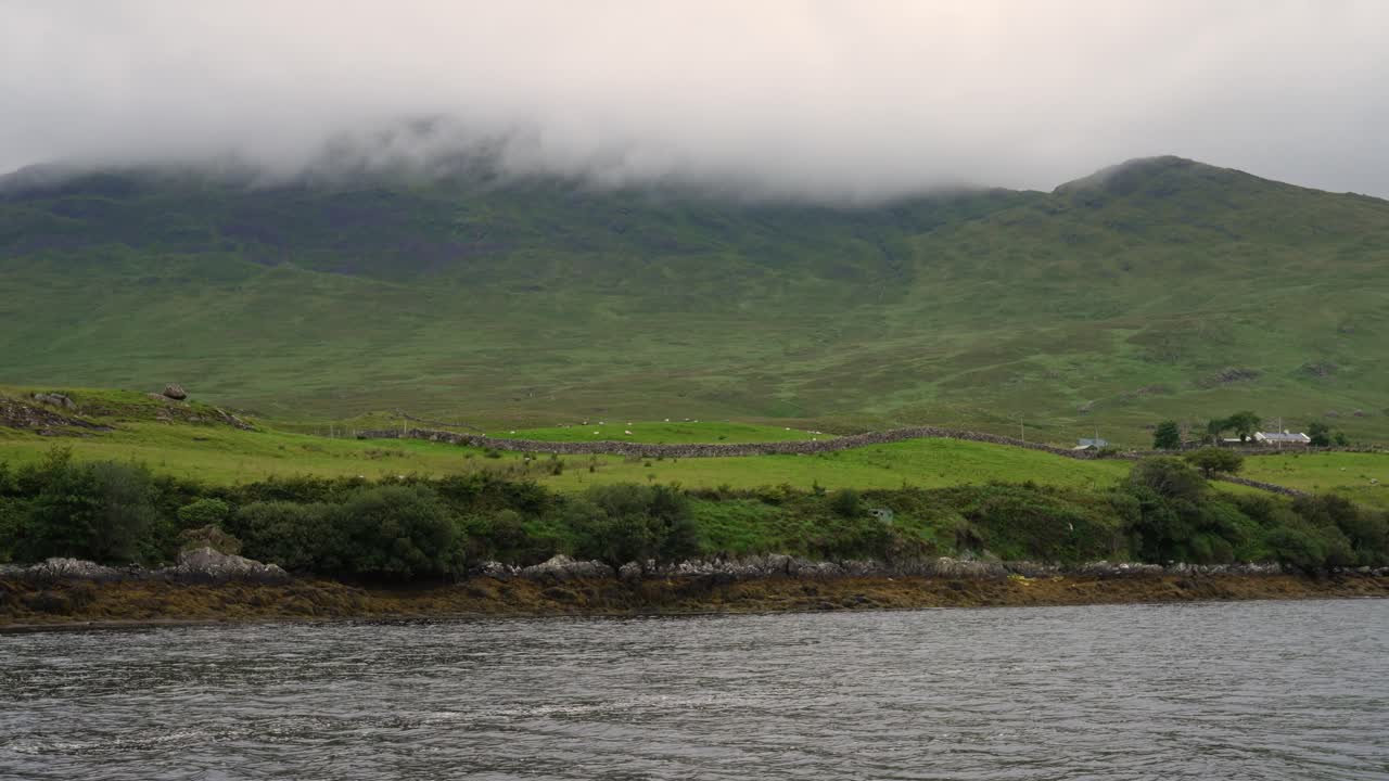 A scenic boat journey along the rugged and misty coastline of Ireland. The view reveals rolling green hills and a remote farmhouse under a cloudy sky