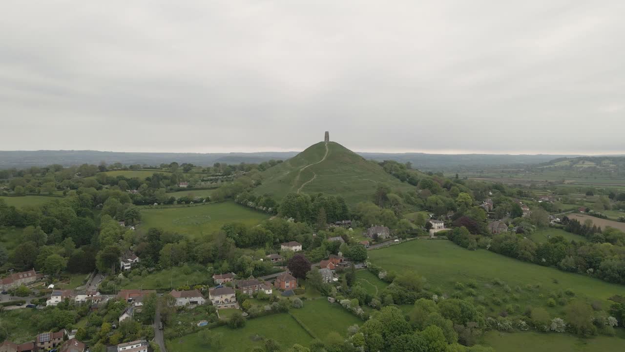 Aerial view of Glastonbury, drone moving backwards moving away from Glastonbury Tor, drone flying over the green fields with some houses on the left side of the camera. 4K, 60fps.