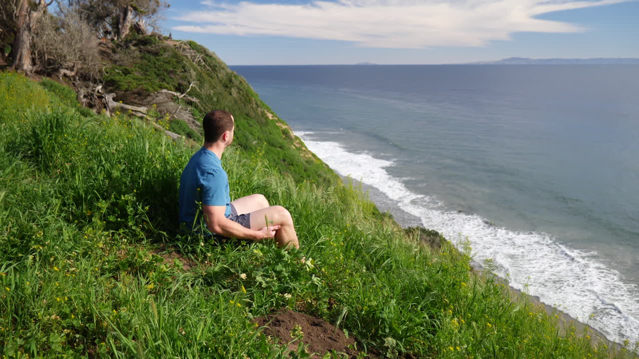 캘리포니아주 산타바바라에서 밝고 화창한 날 깊은 생각과 명상으로 해변 절벽 가장자리에 평화롭게 앉아 있는 남자