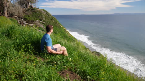 un hombre sentado en paz calmante al borde de un acantilado de playa en pensamiento profundo y meditación en un día soleado en santa barbara, california cámara lenta