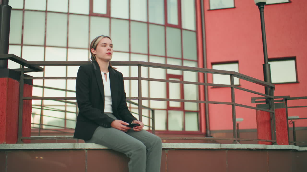 Lady holding phone seated on fence in urban area with striking modern building behind, metal railings around, calm expression, natural light, professional attire, reflective mood in outdoor setting