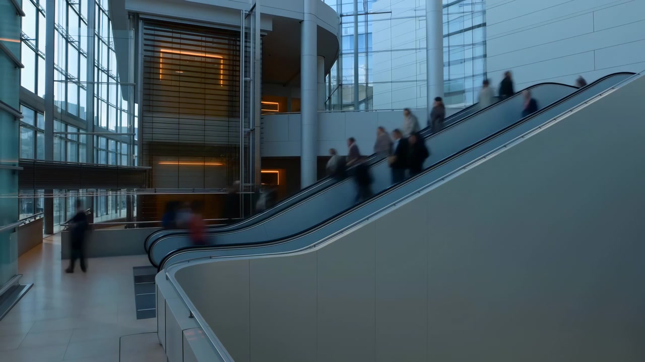 Passageiros embaçados sobem em escadas rolantes paralelas dentro de um átrio multinível iluminado, criando um fluxo constante, ritmo diagonal, reflexos e uma circulação urbana proposital, com a escada rolante e os passageiros