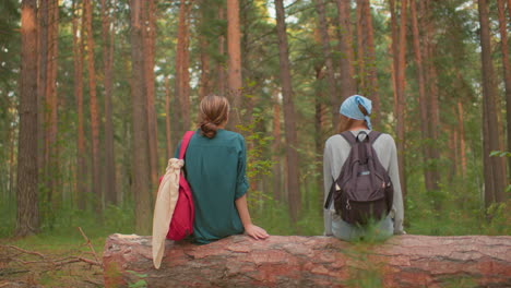 hikers share warm moment on fallen tree in peaceful forest, one wearing blue bandana and black backpack smiles warmly, while her companion in green shirt with red bag cleans her hand after sitting