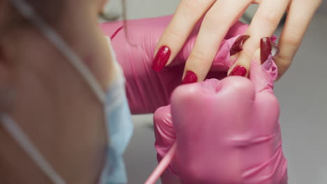 close up of nail technician wearing mask and pink glove carefully applying red nail polish using tiny brush to refine edge of client nail, demonstrating precision and professional beauty care focus