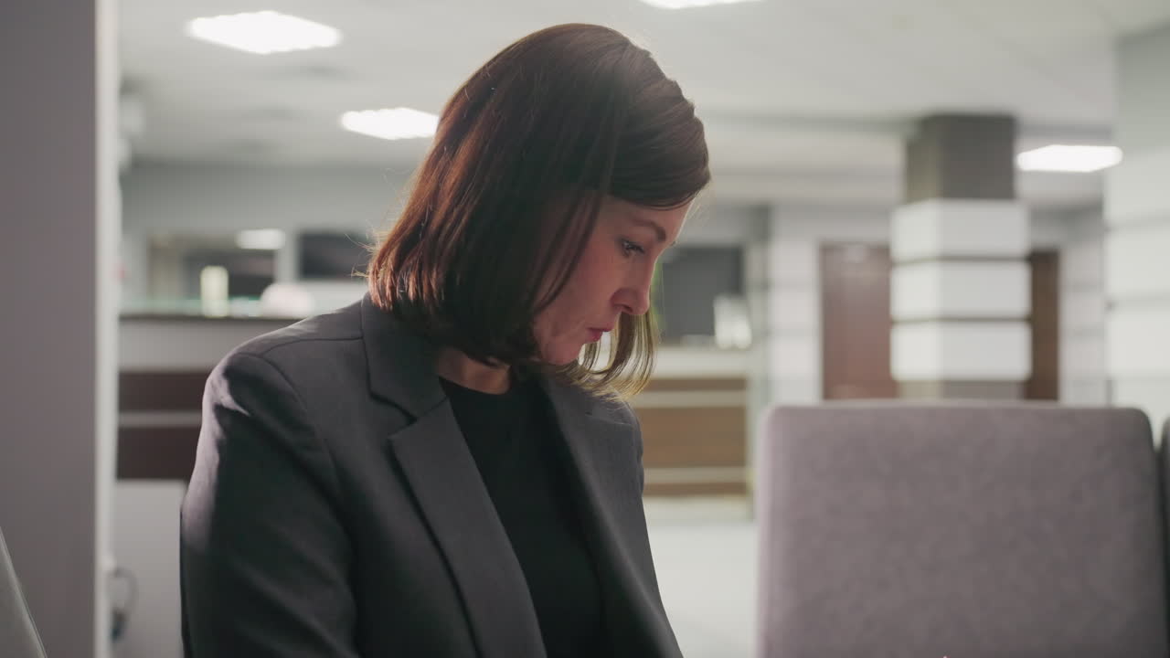 Close-up of businesswoman focused on work, looking down and writing in notebook in modern office lounge. Female professional exhibiting concentration and thoughtfulness in productive office space