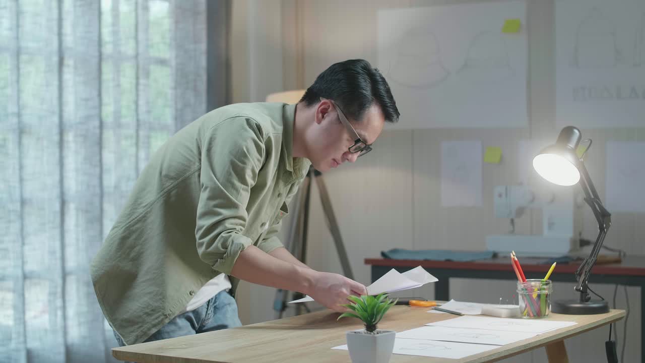 아시아 남자 디자이너의 측면 뷰는 사무실의 테이블에 레이아웃을 배치하고 있습니다.