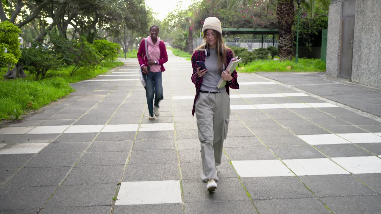 Diversas estudiantes universitarias caminando por el camino del campus, con un teléfono, una amiga las ve, aceleran el paso para abrazarse y hablar