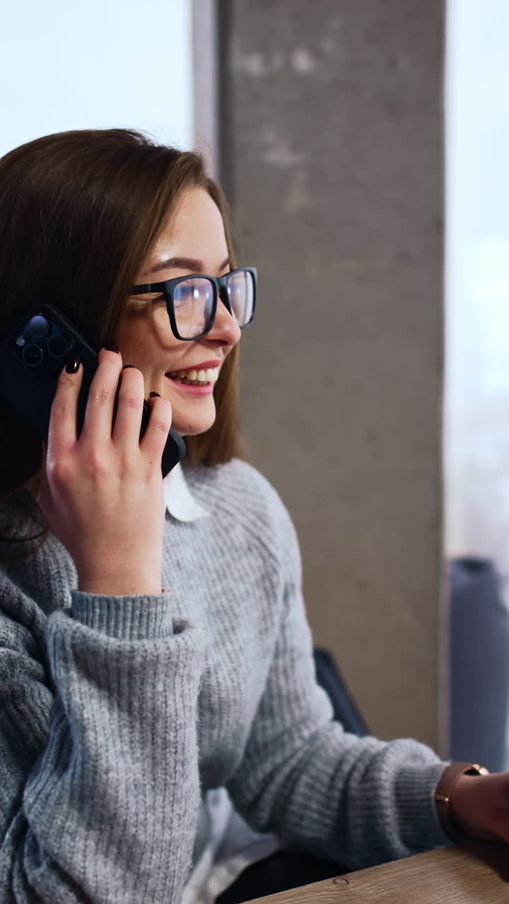 Woman wearing eyeglasses sits at desk speaking on her smartphone. Lady holding a paper cup laughs talking on phone. Vertical video