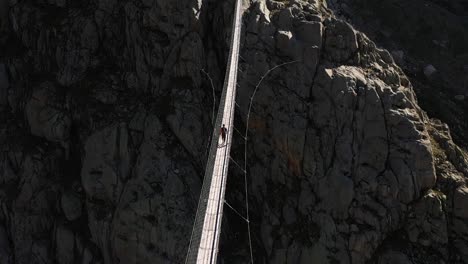 a slow tilting up shot of a single, young man walking confidently alone in the outdoors over a suspension bridge in trifthutte, switzerland