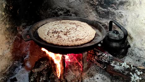 close-up of cooking bread in a cast iron skillet resting on trivets, over an oak wood fire in a large stone fireplace in a traditional kitchen-4