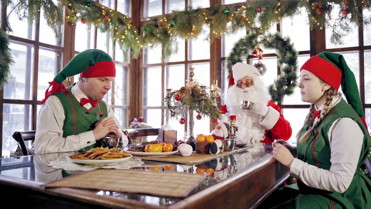 Two funny elves and Santa sitting at festive table on Christmas background. Girl elf tries to take sweets and another elf interferes her.