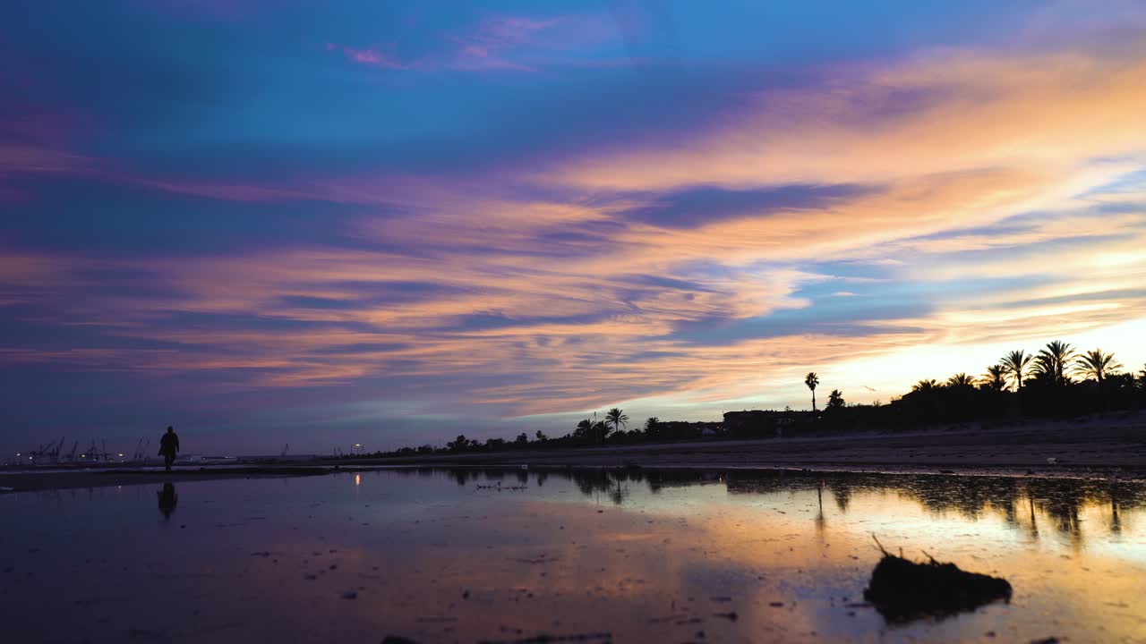 A man walks away calmly walking along the beach in a beautiful sunset, his silhouette is reflected on the water of a lake while he is increasingly walking towards the horizon