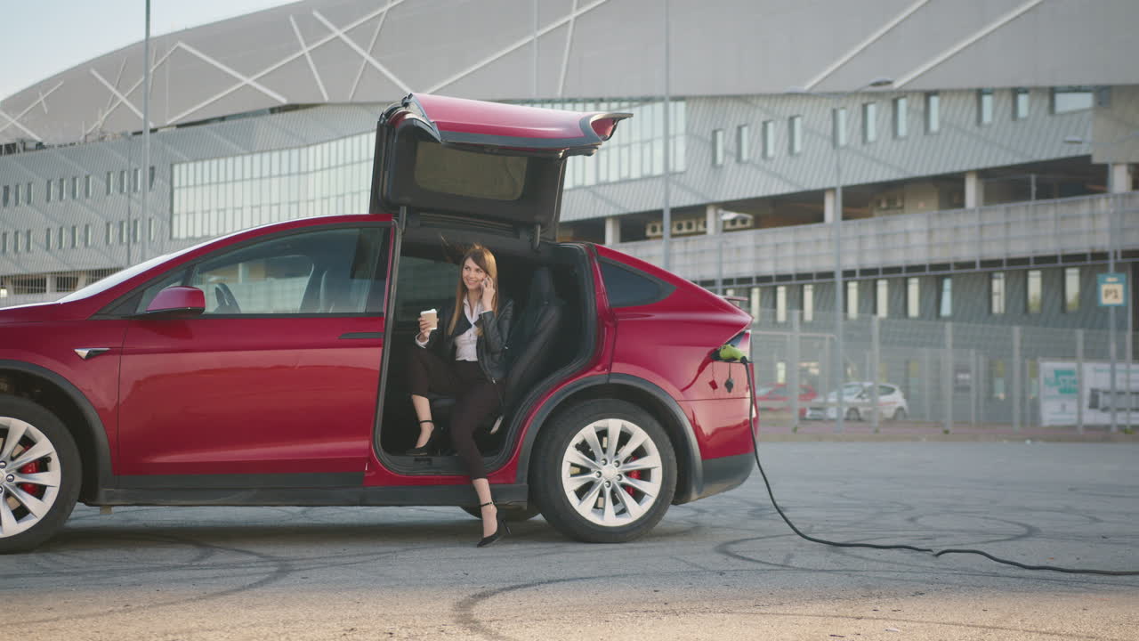 женщина заряжает электромобиль и разговаривает по телефону