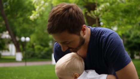 el padre está jugando con su pequeño hijo rubio al aire libre. lo sostiene en sus brazos, jugando. luego lo besa y lo pone en el suelo. hierba verde. cámara lenta