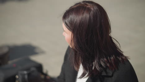 close-up of lady seated, focused on something, illuminated by sunlight, hair swaying in wind, looking away, an unclear object sits close to her