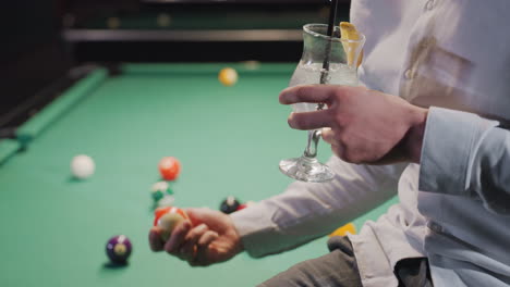 young man in white shirt and grey trousers holding lemon drink with black straw, sitting on green pool table with colorful billiard balls as he rolls a ball. stylish setting, casual mood ambiance