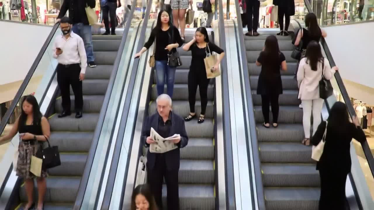 Crowds of shoppers are seen using escalators in a vibrant mall. Some visitors read, while others engage with their mobile devices, showcasing the lively atmosphere of the shopping district.