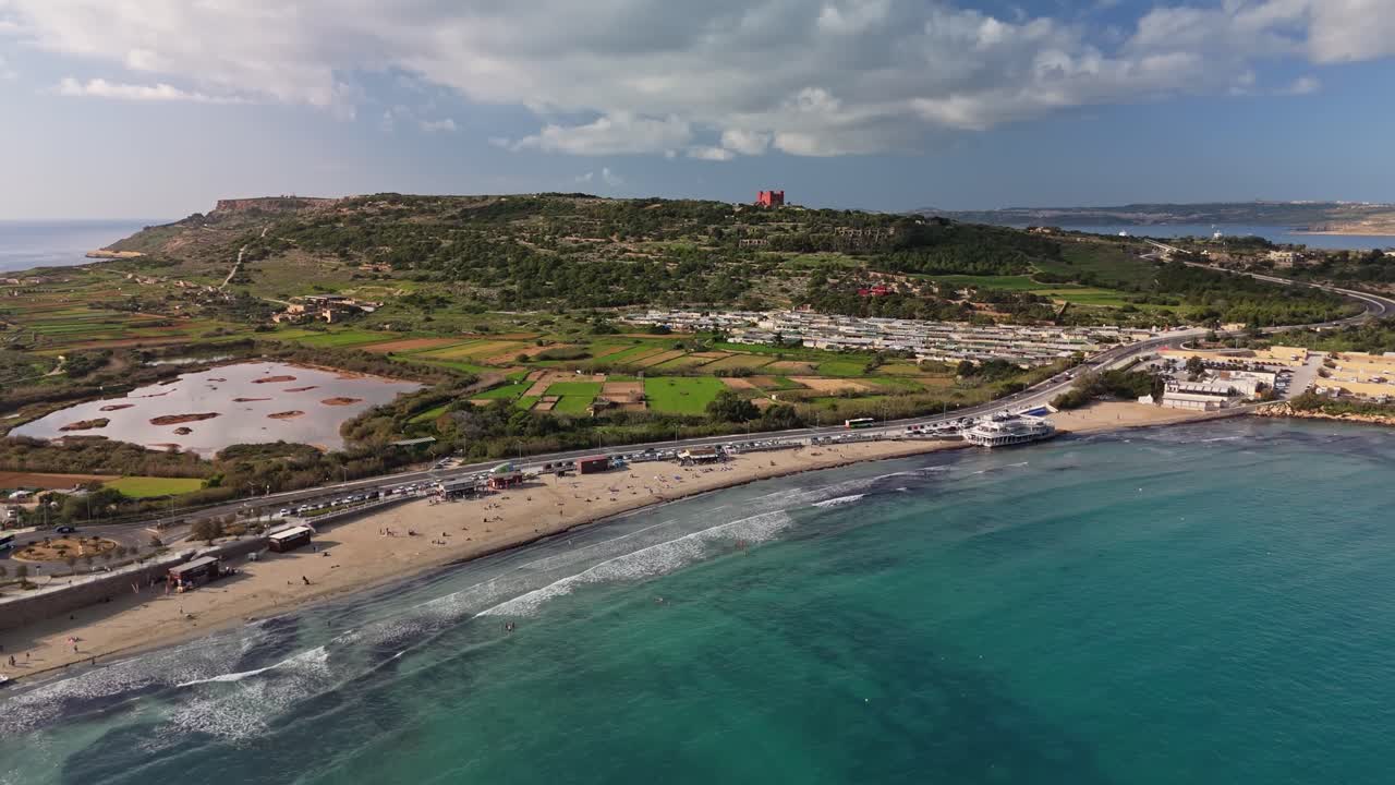 Ripresa aerea in allontanamento di Mellieha, Malta, che mette in risalto la pittoresca baia di Ghadira, la Torre Rossa, le spiagge sabbiose, la riserva naturale con lago e i rigogliosi paesaggi mediterranei sotto un cielo azzurro e nuvoloso