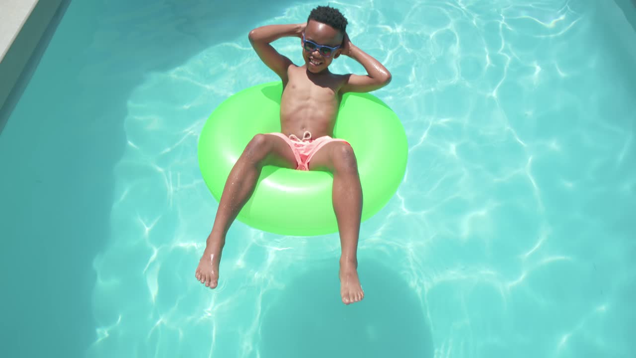Drone rising, teen relaxing on green tube drifting in home pool in pink trunks adjusting sunglasses