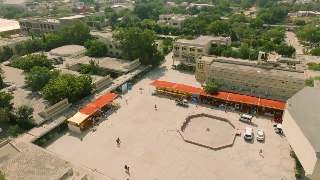 Drone receding and ascending over bustling central open yard of NED University, showing people walking, trees, campus buildings, and the illuminated Karachi city skyline in the background