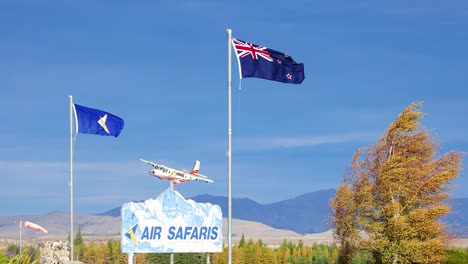 a single-engine aircraft ascends past waving flags and an 'air safaris' sign at a rural airport, with clear skies and mountain backdrop