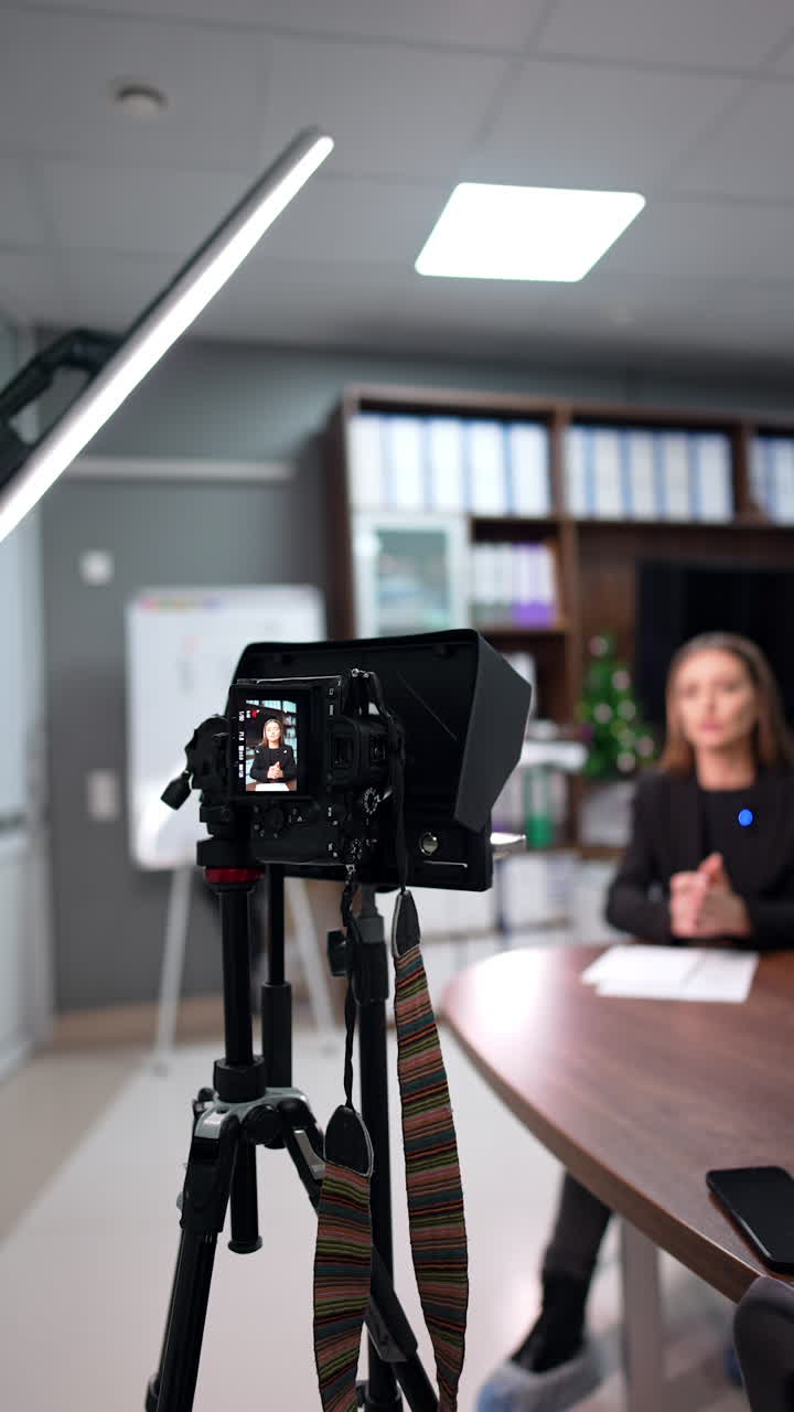 Camera set on a tripod near a lamp to film a lady speaker. Blogger in black jacket talks sitting at desk. Blurred backdrop. Vertical video.