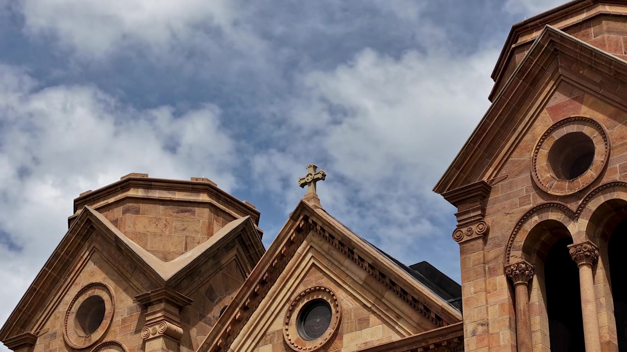 Un drammatico timelapse meteorologico di nuvole in rapido movimento che si spostano dietro un crocifisso in cima a una cattedrale gotica. Il cielo dinamico crea uno sfondo suggestivo contro la storica architettura religiosa