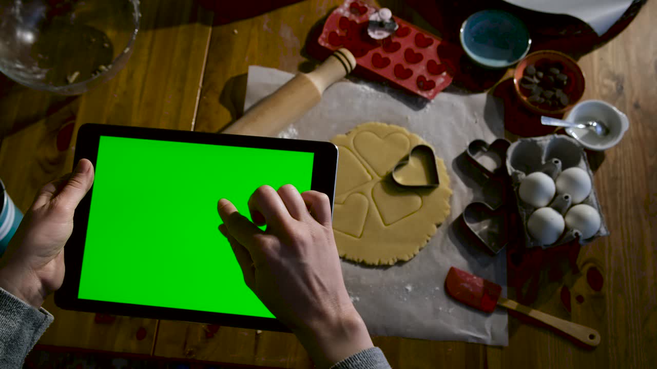 mujer con la mano mirando la receta o las noticias de la tableta en la cocina mientras hace galletas. tableta de pantalla verde.