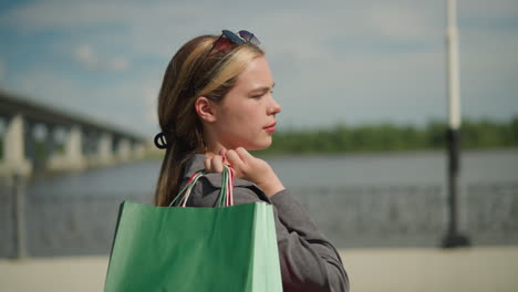 lady carries shopping bag backward as she gazes into the distance, the background features a blur of a couple walking near the road edge, the woman flaunting her hair, and a distant bridge