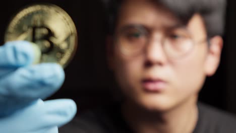 young guy holding bitcoin coin crypto currency in hand with blue gloves and black background