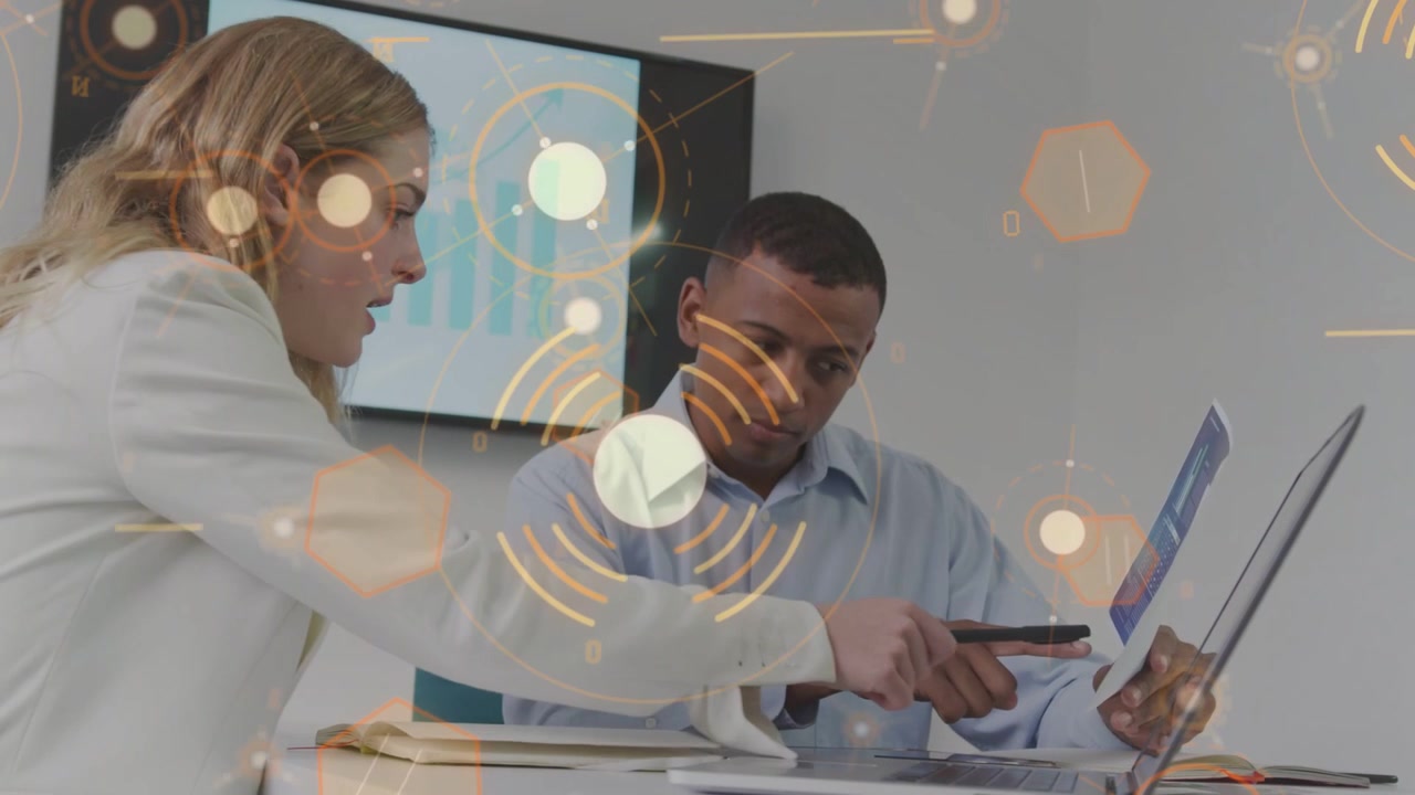 Man pointing brochure at laptop starting animated HUD and woman noting both reviewing business data