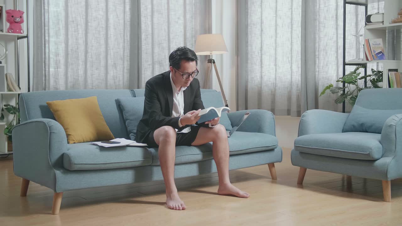 집에서 노트북으로 일한 후 책을 읽는 재과 쇼트를 입은 아시아 사업가의 전체 시각