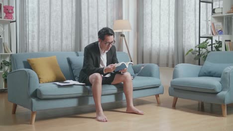 full view of asian businessman in jacket and shorts reading a book after working with a laptop at home