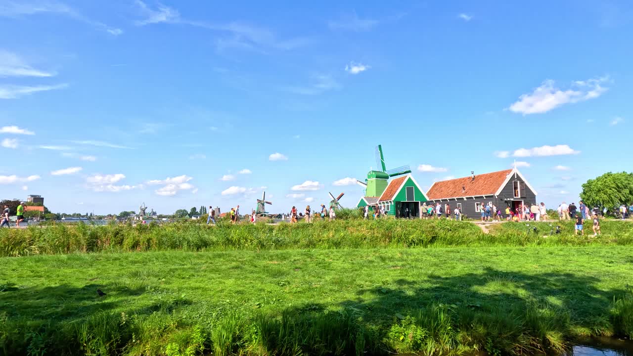 Daytime camera pan reveals windmills, canal, green fields, traditional houses, and tourists in Zaandam