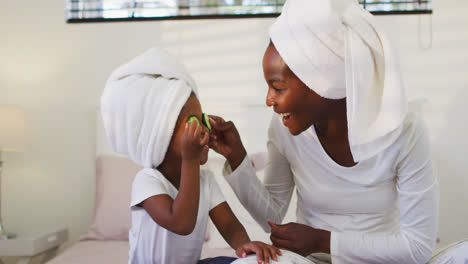 Happy-african-american-mother-and-daughter-wearing-towels-sitting-on-bed-putting-cucumbers-on-eyes