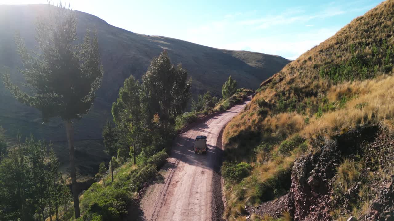Aerial 4K drone footage following a tuk-tuk winding through the golden Peruvian Andes. Warm evening light bathes the mountains in rich colour and texture