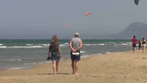 Paar-Mittleren-Alters-Spaziert-Am-Sandstrand-Mit-Kitesurfern-Im-Hintergrund,-Totalaufnahme