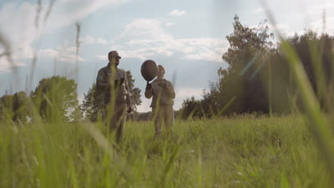couple in matching flight suits preparing tandem glider launch over sunlit grassy field under blue sky breeze, checking harness cords, inspecting canopy inflation